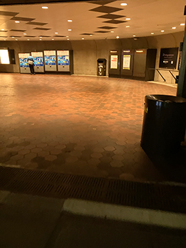 A picture of a pathway in a metro station. In the foreground to the right there is a trash can. In the background there is a wall with fare vending machines slightly to the right of center as well as to the left. In the far right background there is the ending of a railing.